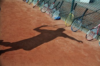 Schatten eines Kindes beim Aufschlag auf rotem Sandplatz, im Hintergrund aufgereihte Tennisschlaeger